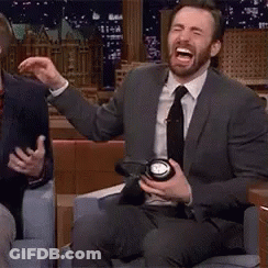 A Man In A Suit And Tie Is Sitting On A Couch Laughing And Holding A Camera . GIF