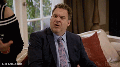 A Man In A Suit And Tie Is Sitting On A Couch With A Woman Behind Him GIF
