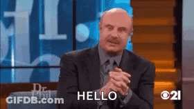 A Man In A Suit And Tie Is Sitting On A Stage With His Hands Folded And Saying Hello . GIF