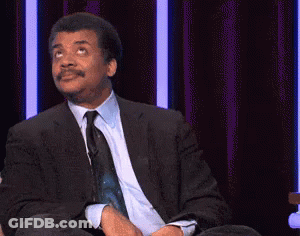 A Man In A Suit And Tie Is Sitting On A Stage With The Words `` Just Kidding '' Written On The Screen . GIF