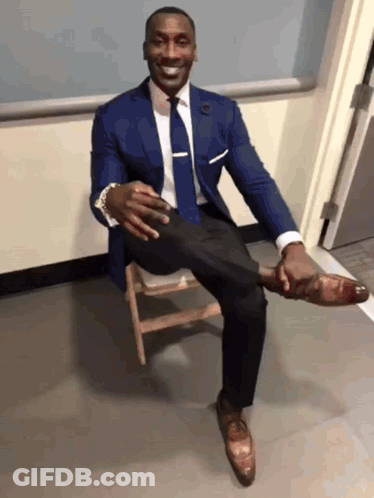 A Man In A Suit And Tie Is Sitting On A Wooden Chair With His Leg Up GIF
