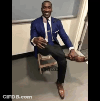 A Man In A Suit And Tie Is Sitting On A Wooden Stool With His Leg Up . GIF