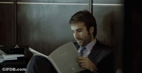 A Man In A Suit And Tie Is Sitting On The Floor Reading A Book GIF