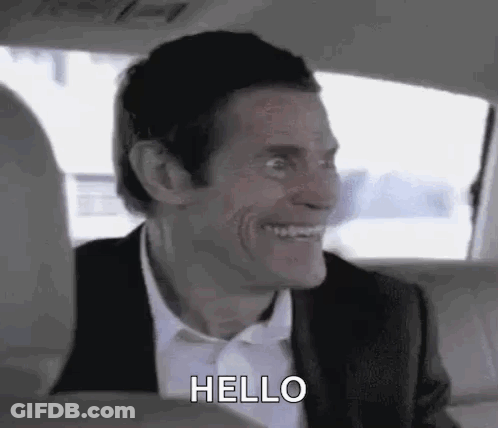 A Man In A Suit And Tie Is Smiling And Saying Hello While Sitting In The Back Seat Of A Car . GIF