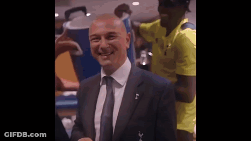 A Man In A Suit And Tie Is Smiling For The Camera While A Man In A Yellow Shirt Stands Behind Him . GIF