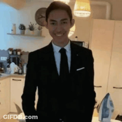 A Man In A Suit And Tie Is Smiling In A Kitchen While Standing Next To An Iron . GIF