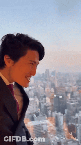 A Man In A Suit And Tie Is Smiling In Front Of A City Skyline GIF