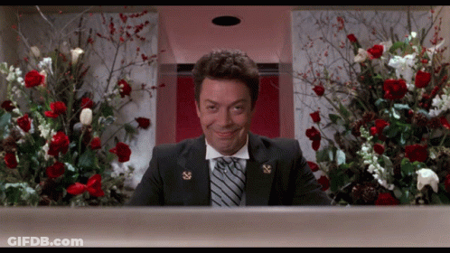 A Man In A Suit And Tie Is Smiling In Front Of A Display Of Flowers GIF