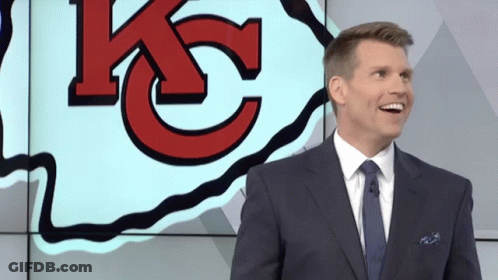 A Man In A Suit And Tie Is Smiling In Front Of A Large Kc Logo GIF