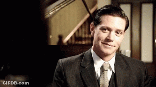 A Man In A Suit And Tie Is Smiling In Front Of A Piano . GIF