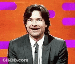 A Man In A Suit And Tie Is Smiling In Front Of A Purple And Orange Background . GIF