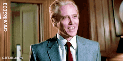 A Man In A Suit And Tie Is Smiling In Front Of A Sign That Says Cravello2023 GIF