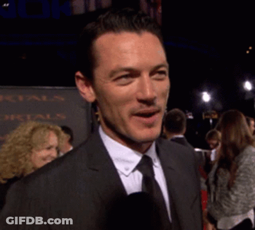 A Man In A Suit And Tie Is Smiling In Front Of A Sign That Says Mortals GIF