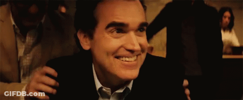 A Man In A Suit And Tie Is Smiling While Being Hugged By Another Man In A Dark Room . GIF