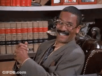 A Man In A Suit And Tie Is Smiling While Sitting In Front Of A Bookshelf . GIF