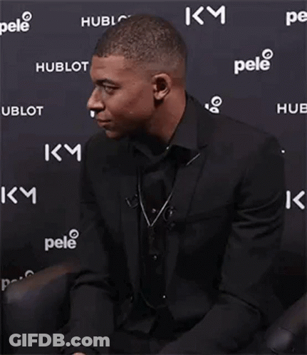 A Man In A Suit And Tie Is Smiling While Sitting In Front Of A Wall With Logos On It . GIF