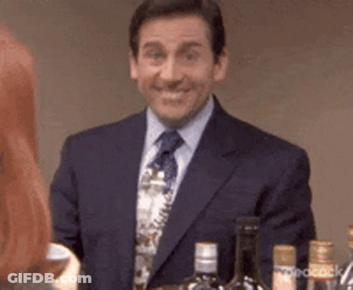 A Man In A Suit And Tie Is Smiling While Standing In Front Of A Bar With Bottles Of Alcohol . GIF