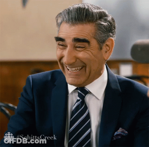 A Man In A Suit And Tie Is Smiling With #Schitts Creek In The Upper Right Corner GIF
