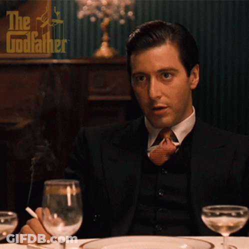 A Man In A Suit And Tie Is Smoking A Cigarette While Sitting At A Table In Front Of A Poster For The Godfather GIF