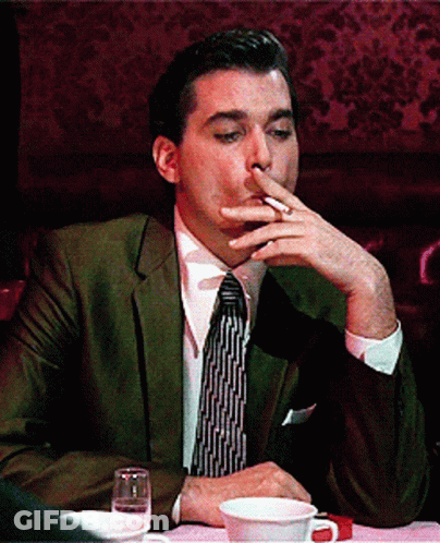 A Man In A Suit And Tie Is Smoking A Cigarette While Sitting At A Table GIF