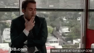 A Man In A Suit And Tie Is Smoking A Cigarette While Sitting In Front Of A Window . GIF