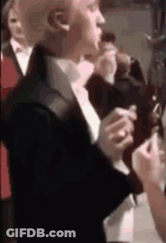 A Man In A Suit And Tie Is Smoking A Cigarette While Standing In A Crowd Of People . GIF