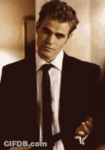 A Man In A Suit And Tie Is Smoking A Cigarette While Standing In A Room . GIF