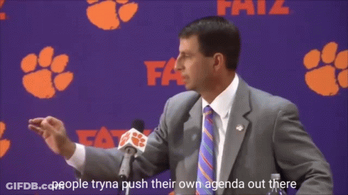 A Man In A Suit And Tie Is Speaking Into A Microphone In Front Of A Clemson Banner GIF