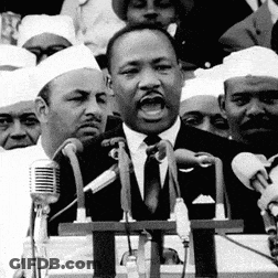 A Man In A Suit And Tie Is Speaking Into A Microphone In Front Of A Crowd Of People . GIF