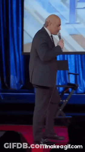 A Man In A Suit And Tie Is Speaking Into A Microphone On A Red Carpet GIF