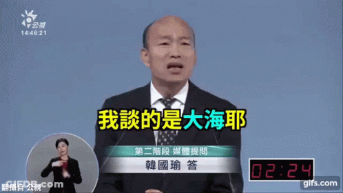 A Man In A Suit And Tie Is Speaking Into A Microphone While A Woman In Sign Language Watches GIF