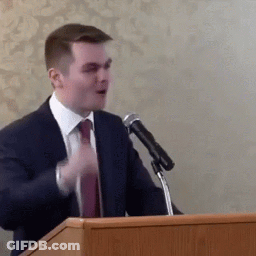 A Man In A Suit And Tie Is Standing At A Podium Giving A Speech In Front Of A Microphone . GIF