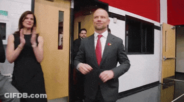 A Man In A Suit And Tie Is Standing In A Doorway With A Woman Clapping Behind Him GIF