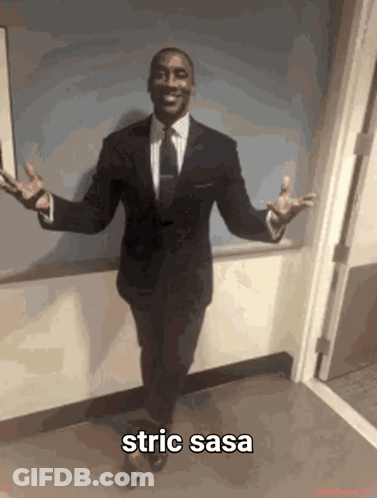 A Man In A Suit And Tie Is Standing In A Hallway With His Arms Outstretched With The Caption Strict Sasa GIF