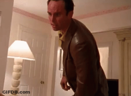 A Man In A Suit And Tie Is Standing In A Room With A Lamp And A Door . GIF