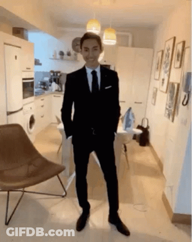 A Man In A Suit And Tie Is Standing In A Room With His Hands In His Pockets . GIF