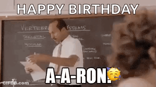 A Man In A Suit And Tie Is Standing In Front Of A Blackboard And Holding A Piece Of Paper . GIF