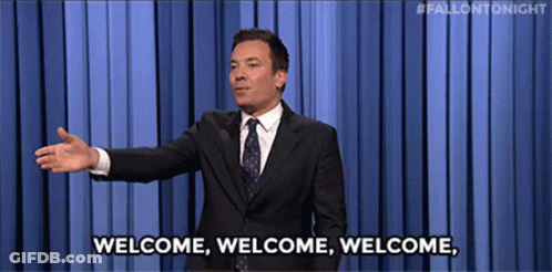 A Man In A Suit And Tie Is Standing In Front Of A Blue Curtain And Says Welcome . GIF