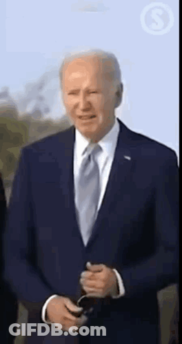 A Man In A Suit And Tie Is Standing In Front Of A Blue Sky And Holding A Cell Phone . GIF