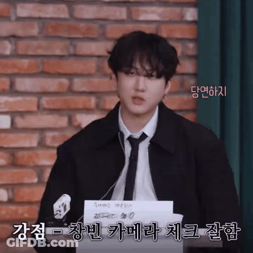A Man In A Suit And Tie Is Standing In Front Of A Brick Wall And Holding A Piece Of Paper With Korean Writing On It GIF