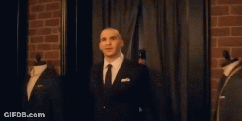 A Man In A Suit And Tie Is Standing In Front Of A Brick Wall In A Room . GIF