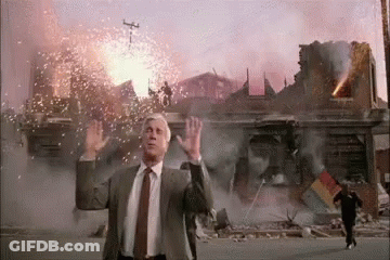 A Man In A Suit And Tie Is Standing In Front Of A Building That Is On Fire . GIF