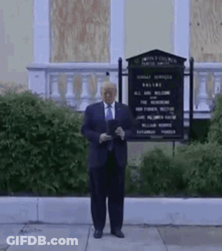 A Man In A Suit And Tie Is Standing In Front Of A Building With A Sign . GIF