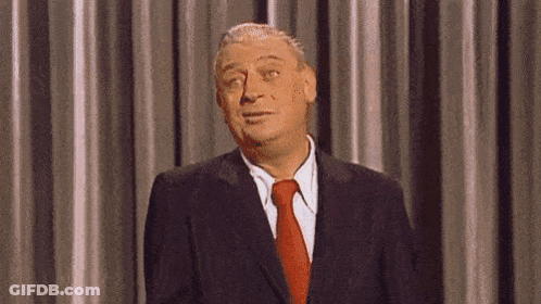 A Man In A Suit And Tie Is Standing In Front Of A Curtain And Making A Funny Face . GIF