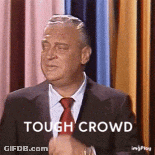 A Man In A Suit And Tie Is Standing In Front Of A Curtain And Says Tough Crowd . GIF