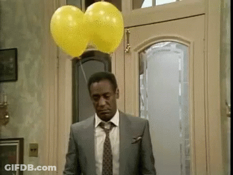 A Man In A Suit And Tie Is Standing In Front Of A Door With Two Yellow Balloons Hanging From It . GIF