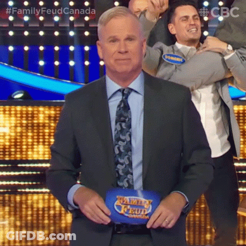 A Man In A Suit And Tie Is Standing In Front Of A Family Feud Podium . GIF