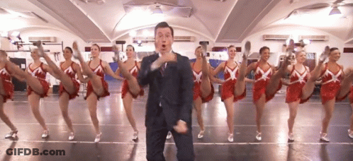 A Man In A Suit And Tie Is Standing In Front Of A Group Of Cheerleaders GIF