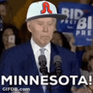 A Man In A Suit And Tie Is Standing In Front Of A Microphone Wearing A Baseball Cap That Says Minnesota . GIF