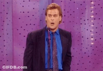 A Man In A Suit And Tie Is Standing In Front Of A Purple Curtain And Pointing At Something . GIF